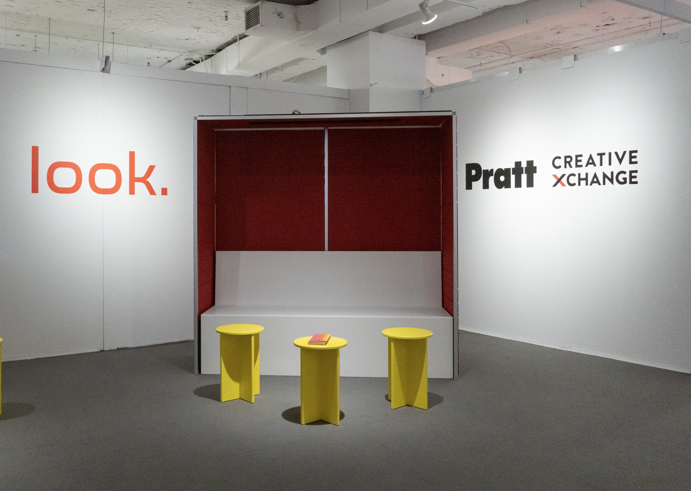 A modern exhibit installation with a bold red and white seating alcove and three yellow stools in front, beneath wall-mounted logos for 